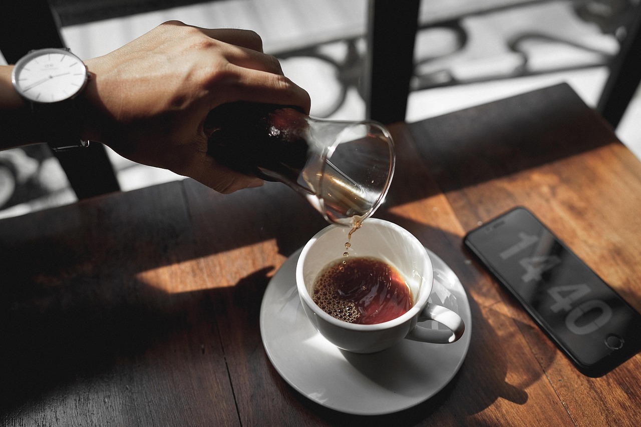 A hand slowly pouring pour-over coffee into a white cup on a wooden table in morning light.