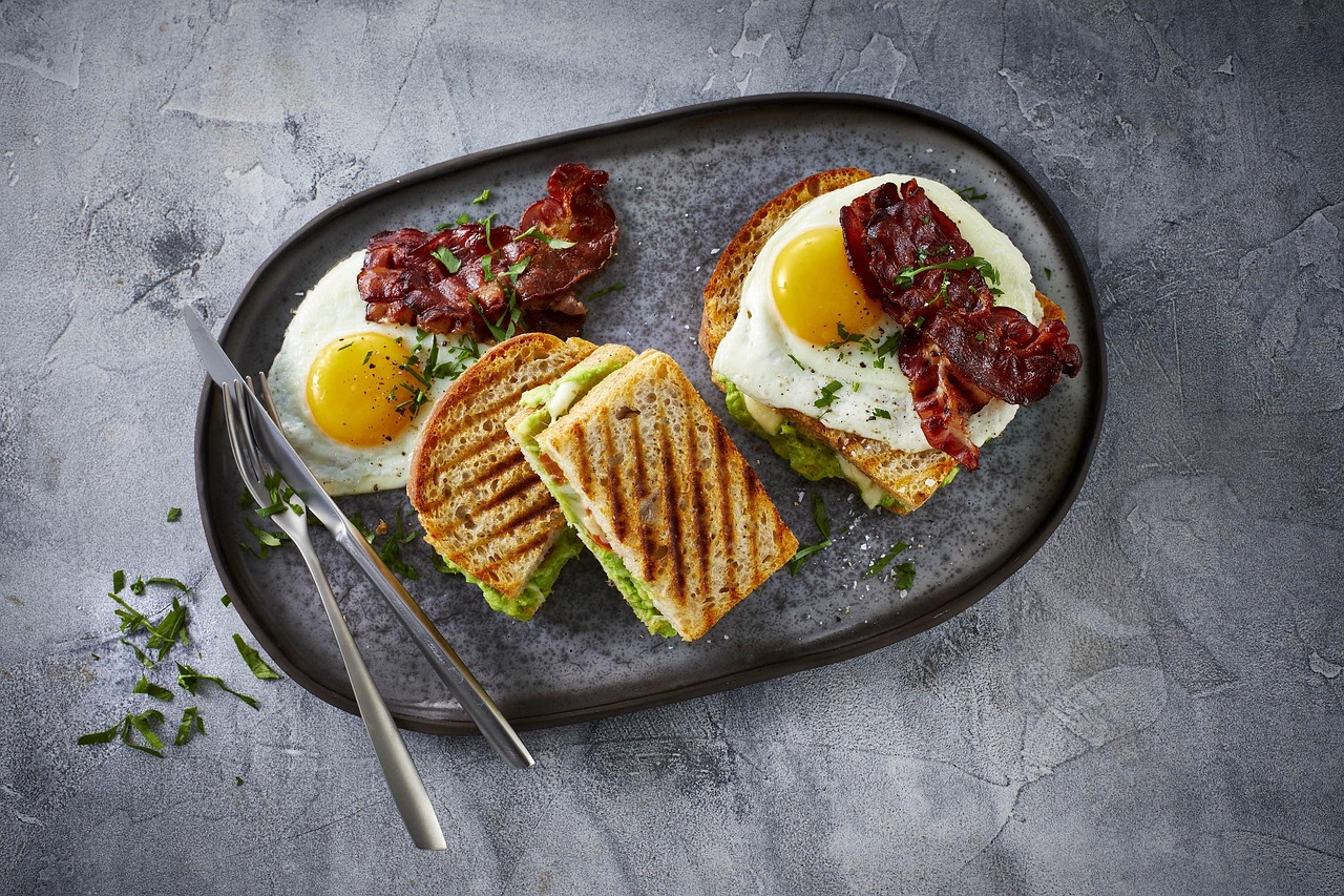 An overhead brunch plate with a fried egg, avocado toast, and crisp bacon on a stone platter.